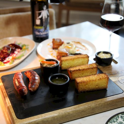 A restaurant board with sausages, grilled bread, salad, dipping sauces, and red wine glasses.