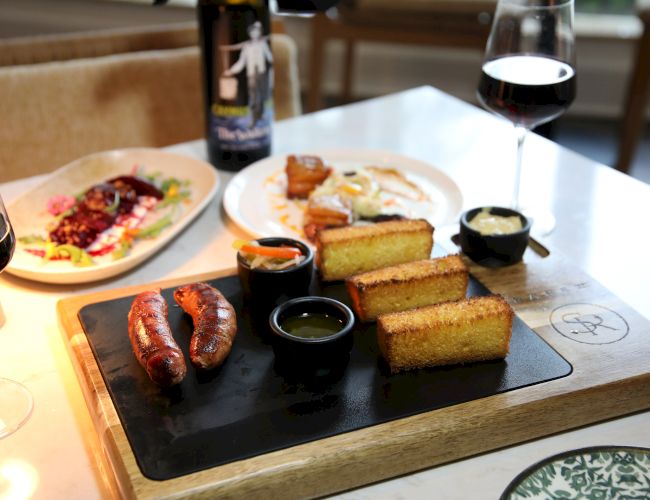 A restaurant board with sausages, grilled bread, salad, dipping sauces, and red wine glasses.