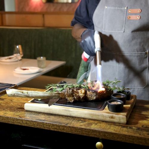 A chef in an apron grills a steak on a wooden board, with rosemary and a small flame during cooking.