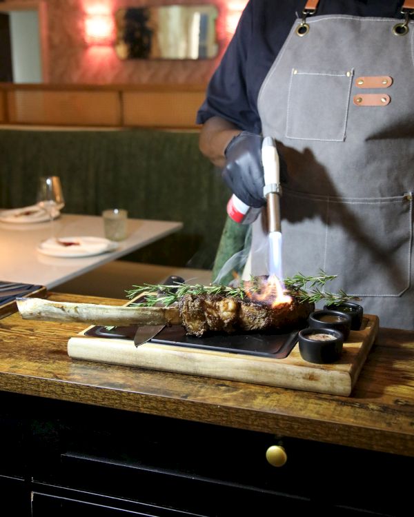 A chef in an apron grills a steak on a wooden board, with rosemary and a small flame during cooking.