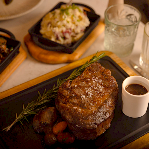 A cooked steak with herbs, a cup of sauce on a tray, and a dish of rice in the background, with glasses and cutlery nearby.