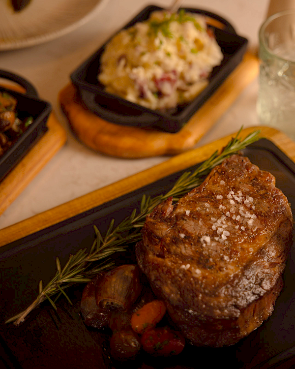 A cooked steak with herbs, a cup of sauce on a tray, and a dish of rice in the background, with glasses and cutlery nearby.