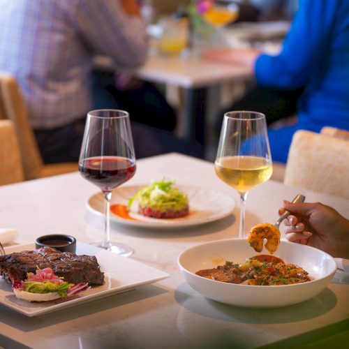 A table with two wine glasses and plates of food, including steak, a salad, and paella/tajine, with people dining in the background.