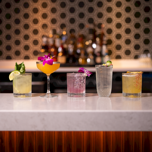 Five colorful cocktails lined up on a glossy bar counter, each with distinct garnishes and flowers, against a dark patterned background.