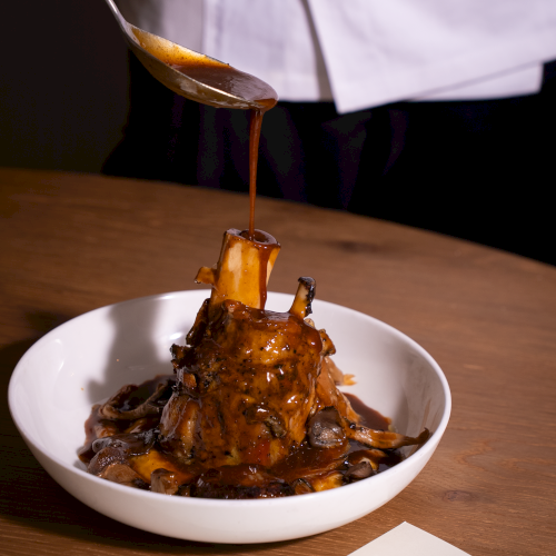 A rich dish with chunky meat and a glossy sauce being poured over it, served in a white bowl.