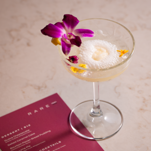 A martini-style cocktail with a frothy top and a purple orchid garnish, placed beside a maroon menu on a marble surface.