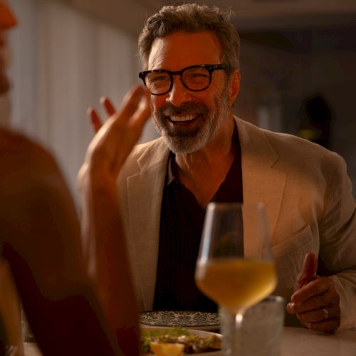 A person with glasses smiles at a dining table, with a glass of wine and food nearby, in a warmly lit setting.